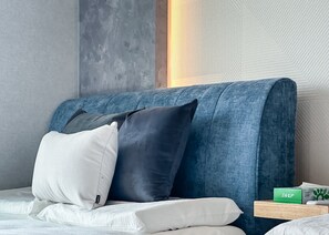 Ein Schlafzimmer mit einem blau gepolsterten Kopfteil, weißer Bettwäsche und einer grünen Schachtel auf einem Nachttisch aus Holz.