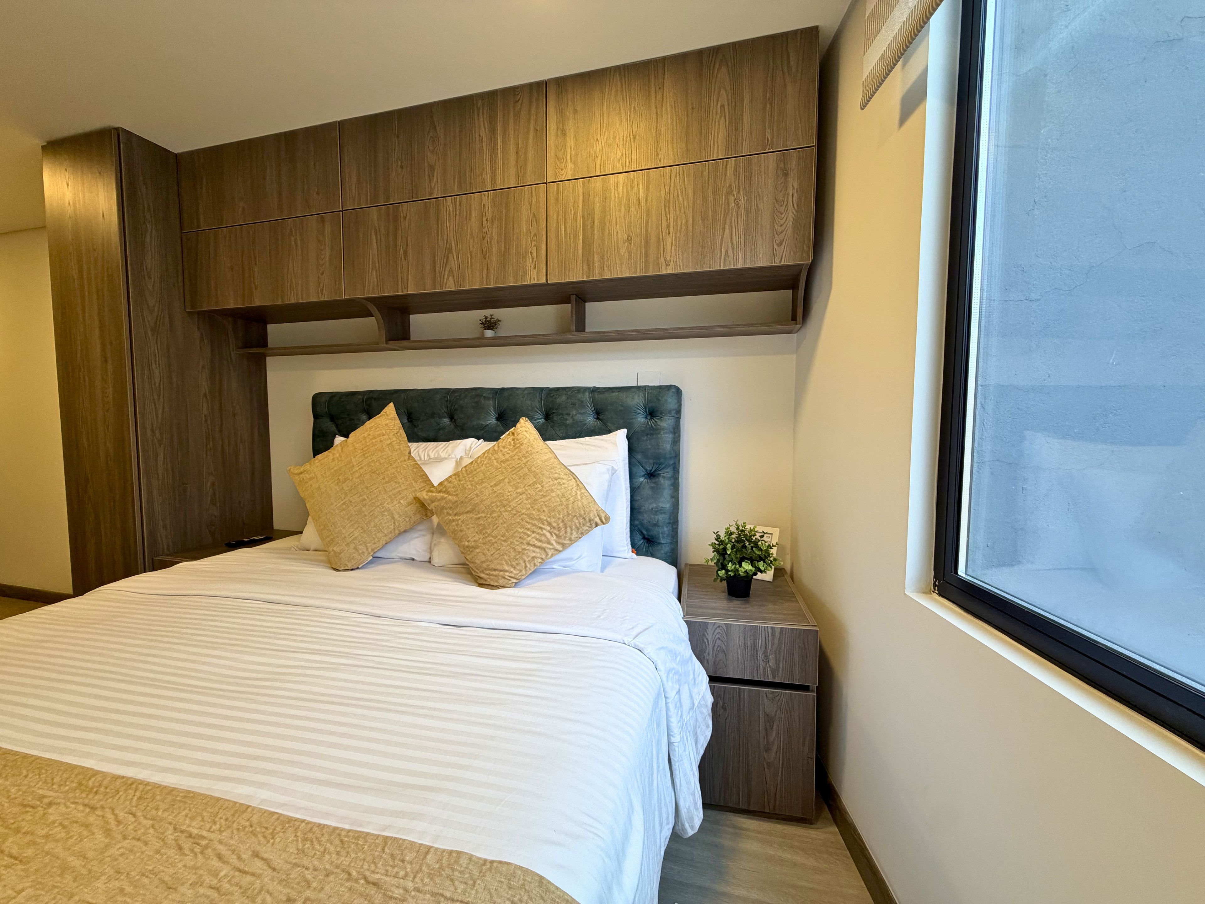 A hotel room with a large bed, wooden headboard, and a small bedside table with a plant.