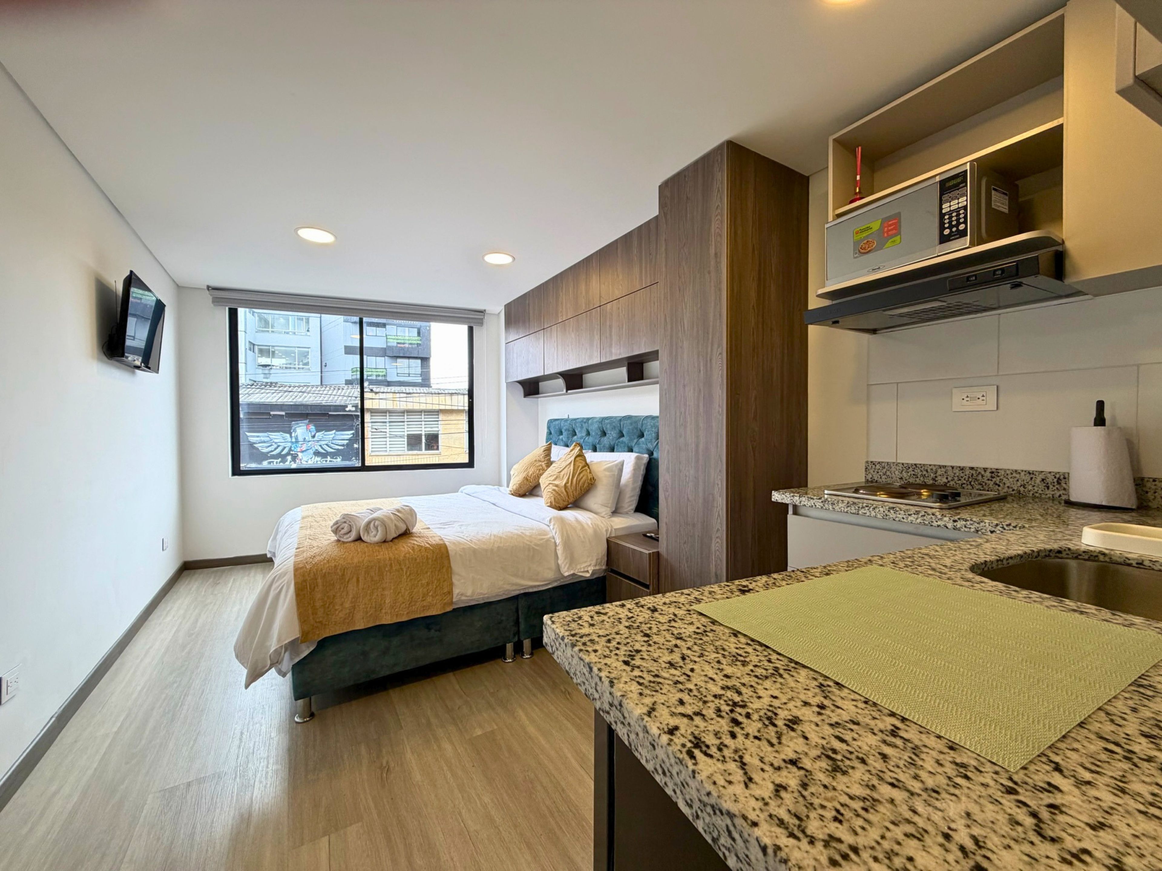 A modern hotel room with a bed, a small kitchenette, a window with a view of buildings, and a television on the wall.