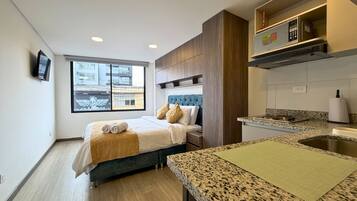 A modern hotel room with a bed, a small kitchenette, a window with a view of buildings, and a television on the wall.