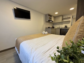 Habitación de hotel con cama, televisor de pantalla plana montado en la pared, kitchenette con microondas y cafetera, y una pequeña planta en maceta.