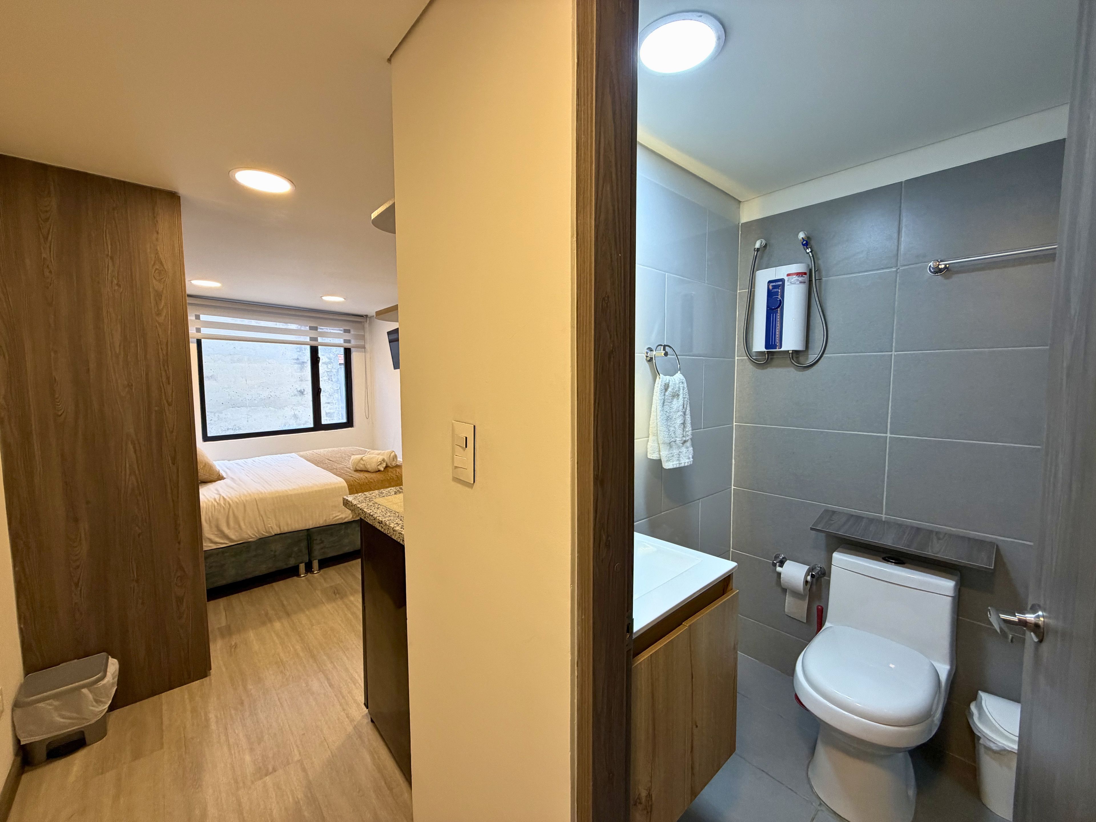 Een moderne badkamer met toilet, douche en een klein wastafeltje. Er is een raam dat voor daglicht zorgt, en een bed in de aangrenzende kamer.