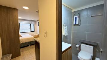 Een moderne badkamer met toilet, douche en een klein wastafeltje. Er is een raam dat voor daglicht zorgt, en een bed in de aangrenzende kamer.