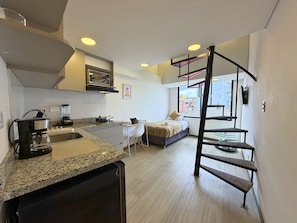 A compact kitchen with a microwave, coffee maker, and blender. There is a bed with a view of the city through a large window. - Nook Galerías by Renteo (Bogotá)