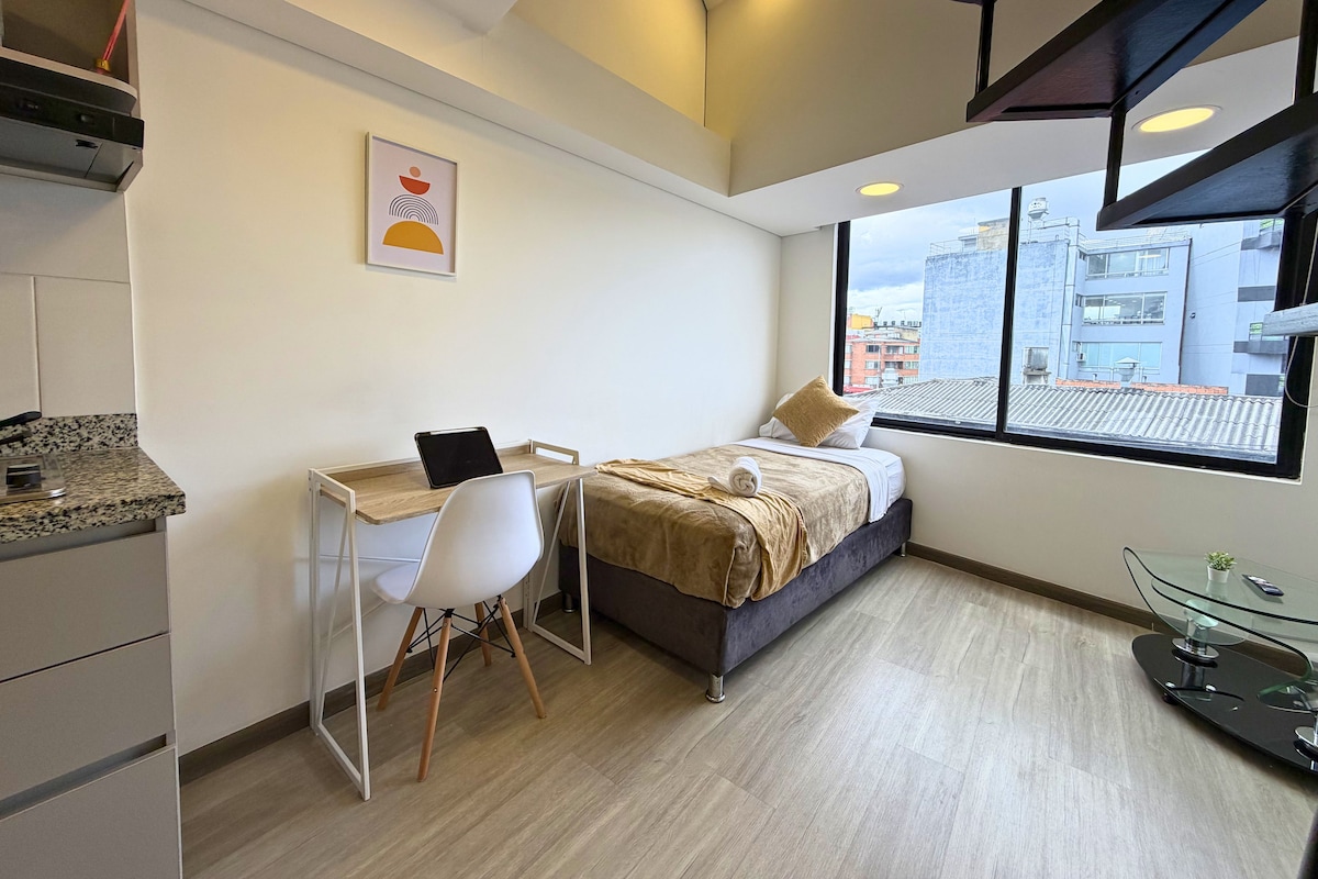 A modern hotel room with a bed, desk, chair, and a view of the city through large windows.