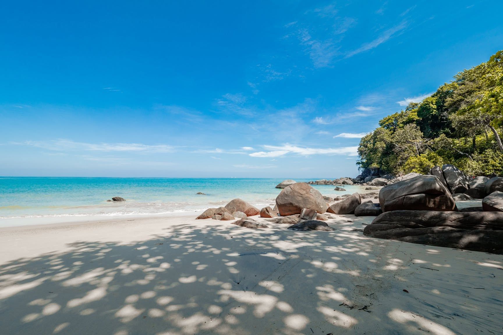 A beach with clear turquoise water, large rocks, and a tree-covered cliff.