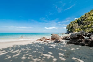 A beach with clear turquoise water, large rocks, and a tree-covered cliff.