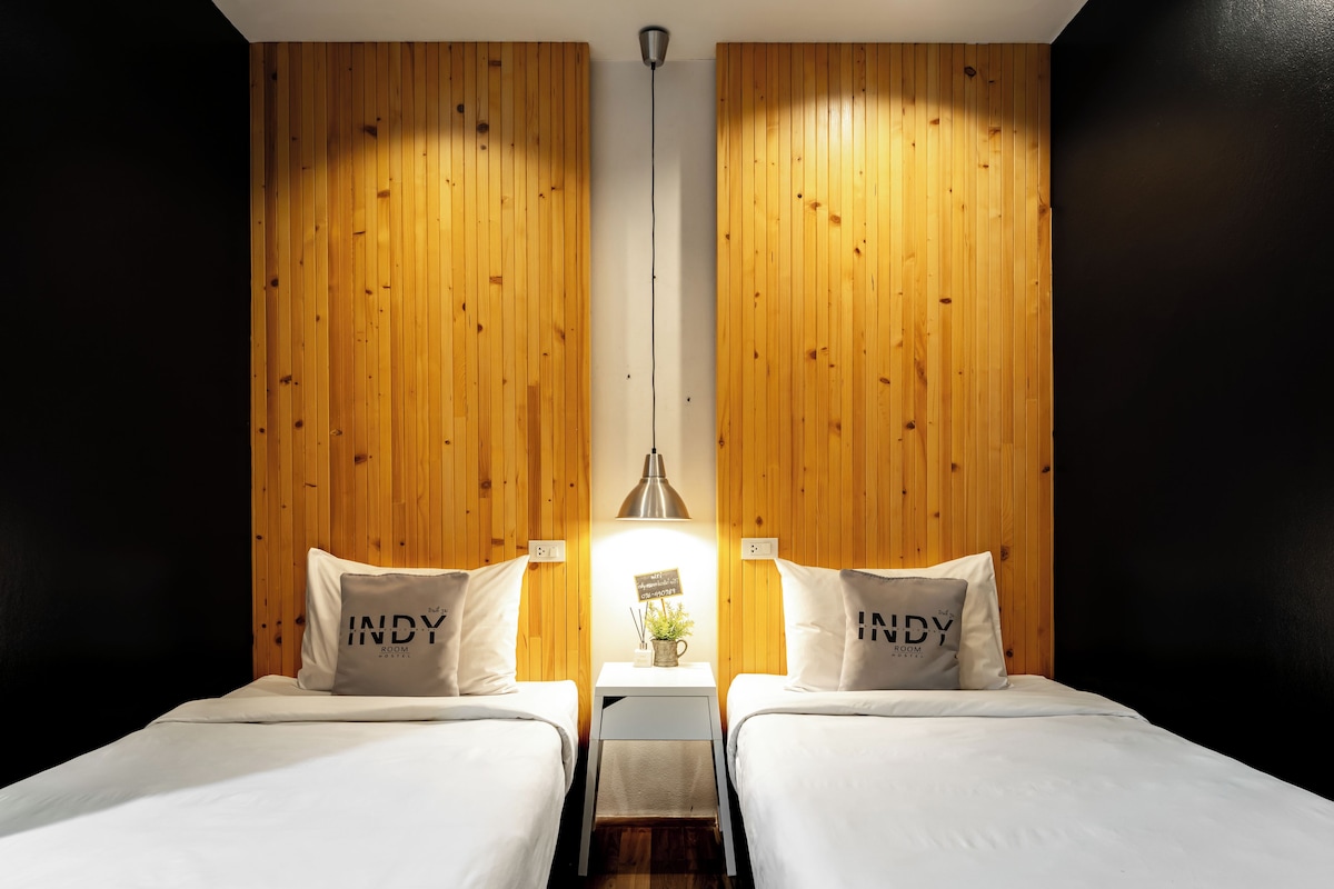 A hotel room with two beds, wooden panel walls, a hanging light fixture, and a small table with a plant.