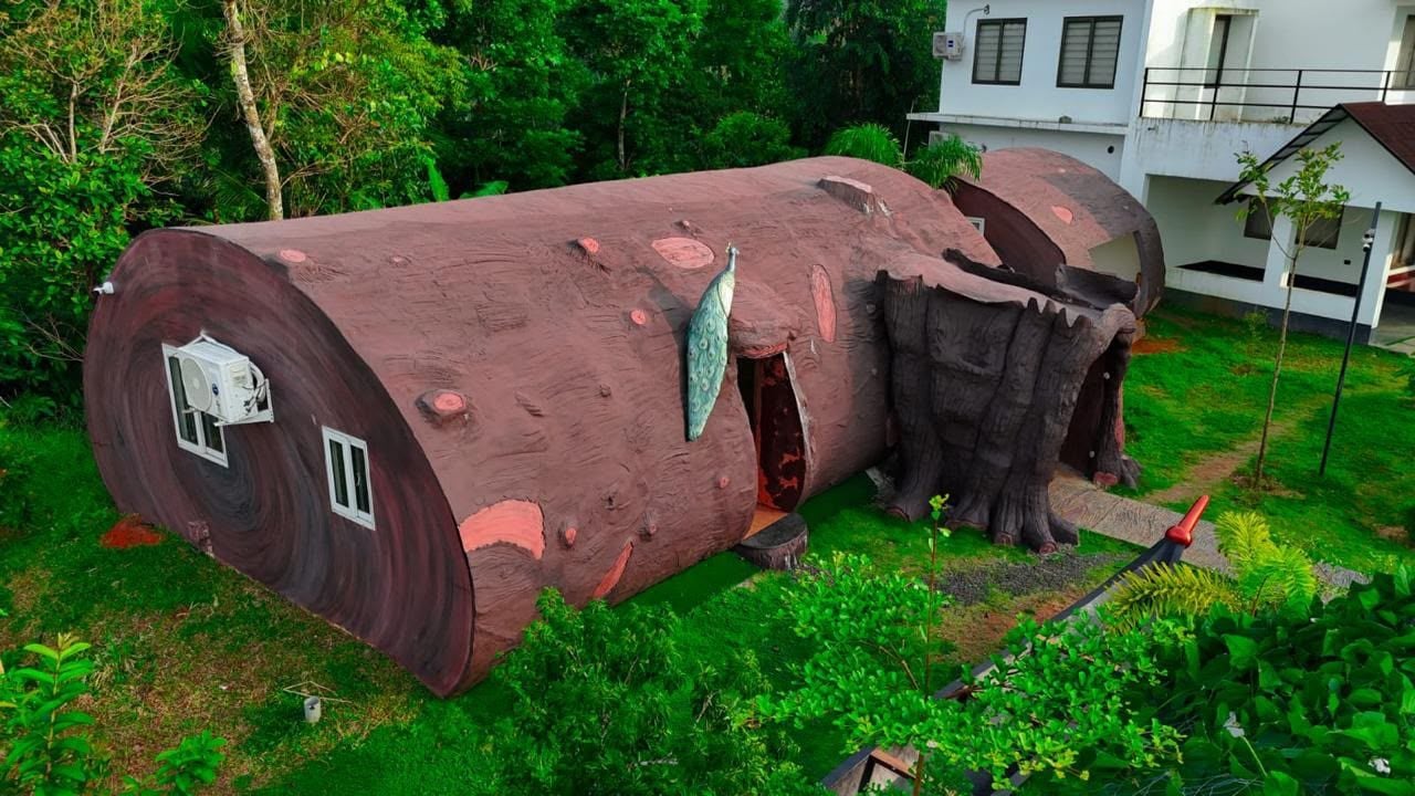 Ein großes Blockhaus mit Fenstern und einer Tür, umgeben von Grünpflanzen und einem Gebäude im Hintergrund.