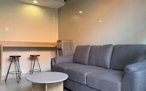 Un espacio de estar moderno con un sofá gris, dos taburetes de madera, una mesa de centro redonda y una máquina de café espresso integrada.