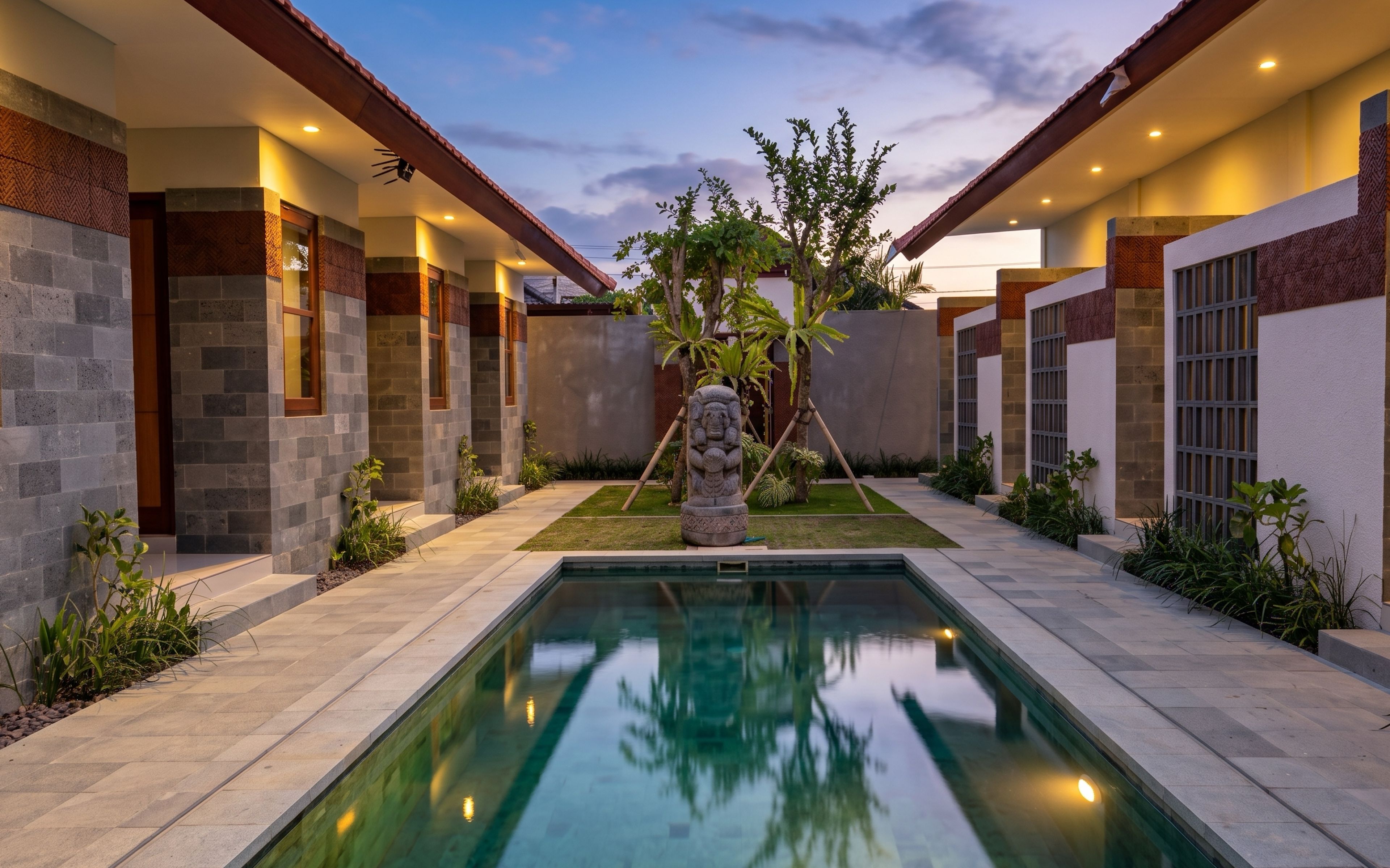 A modern pool area with a stone sculpture, surrounded by a well-maintained garden and a building with a herringbone pattern on the walls.