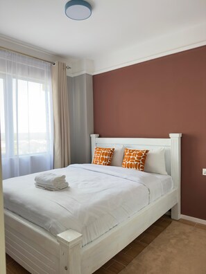 A neatly made bed with white bedding and orange patterned pillows, a white headboard, and a beige carpet. - TALANTA SKYLINE ONE BEDROOM ALONG NGONG ROAD, NEAR JUNCTION MALL (Nairobi)
