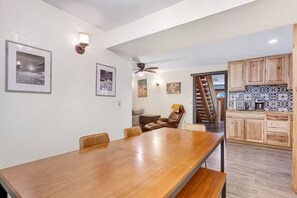 Een eetgedeelte met een lange houten tafel en stoelen, een plafondventilator, en een keuken met houten kasten en een betegelde spatwand.