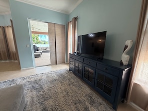 Een woonkamer met een flatscreen televisie op een donkere kast, een vloerkleed met dessin en een uitzicht door een schuifglazen deur naar een buitengebied.