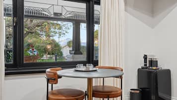A room with a round table, two chairs, a window with a view of a bridge and greenery, and a small black cabinet with a coffee machine.