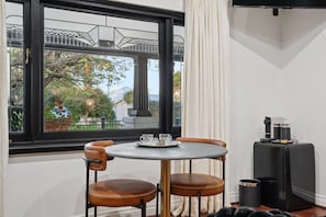 A room with a round table, two chairs, a window with a view of a bridge and greenery, and a small black cabinet with a coffee machine.