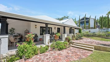 Una casa de dos pisos con un patio de ladrillo, mobiliario exterior y un jardín con diversas plantas y flores.