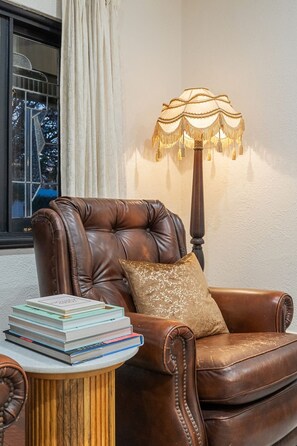Un sillón de cuero con una pila de libros, una lámpara y una ventana con cortinas.