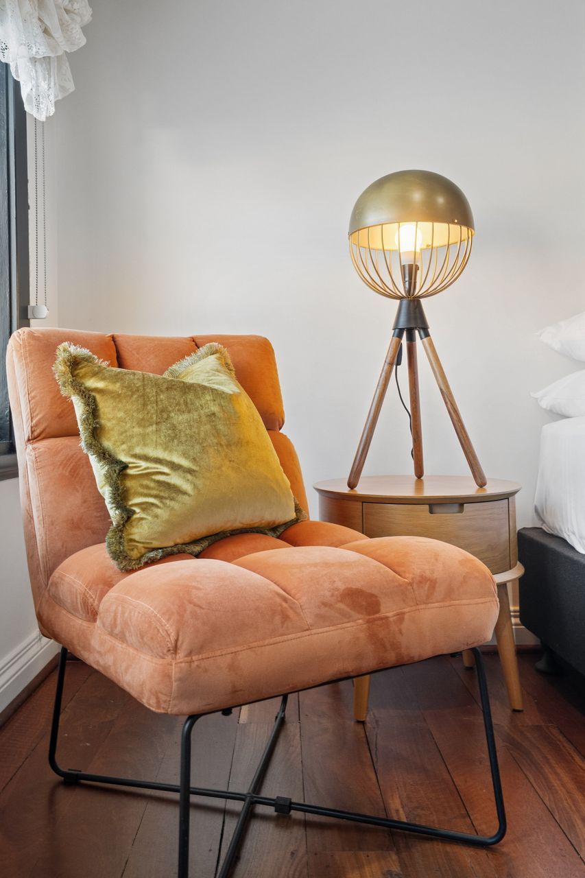 A room with a single orange chair, a yellow pillow, and a floor lamp with a tripod base.