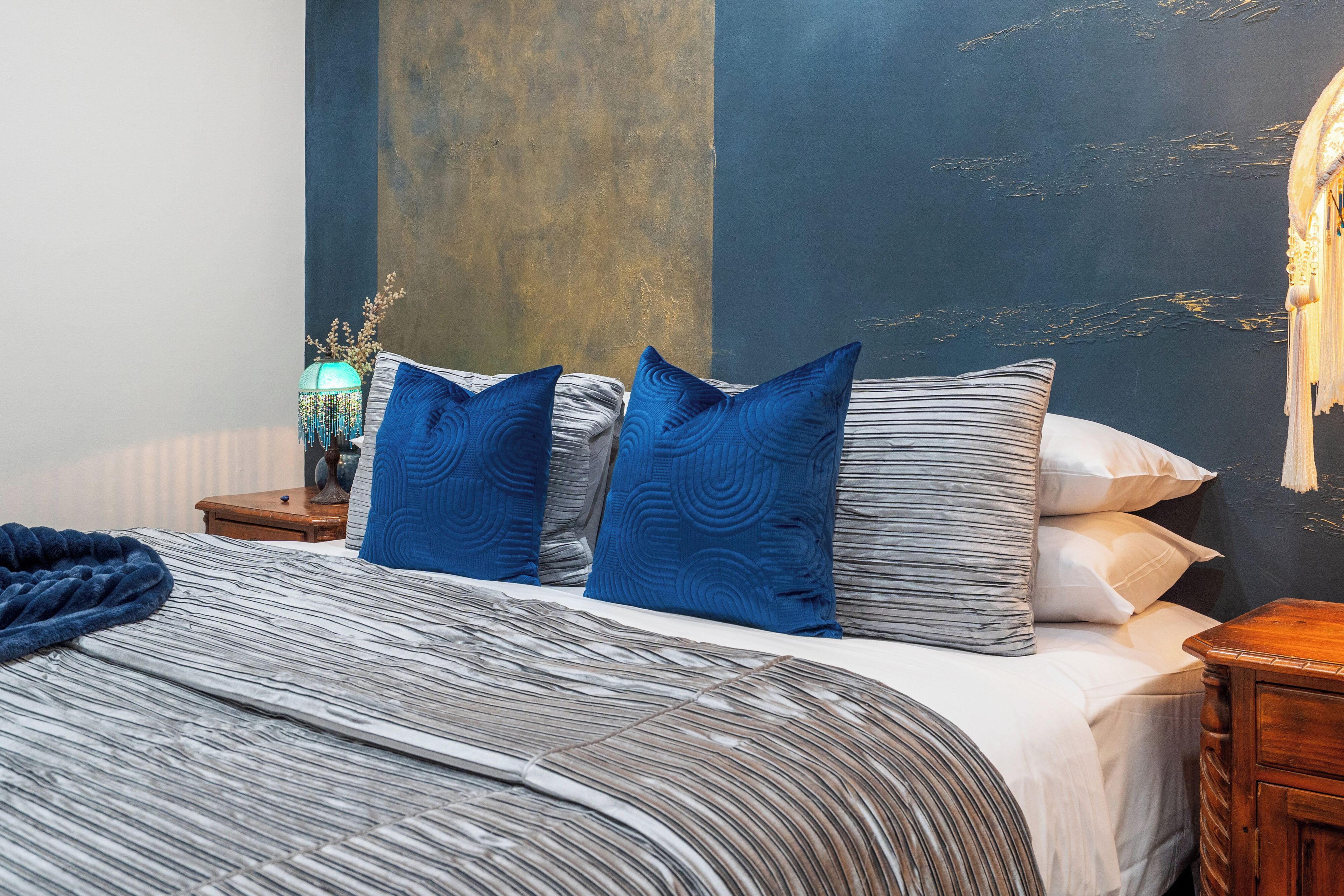 A bed with a striped blanket and blue pillows, a bedside table with a lamp, and a wall with a textured finish.