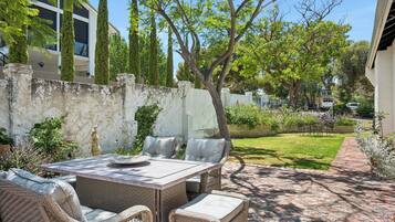 Un patio al aire libre con una mesa y sillas de mimbre, rodeado de follaje y un sendero de ladrillo.