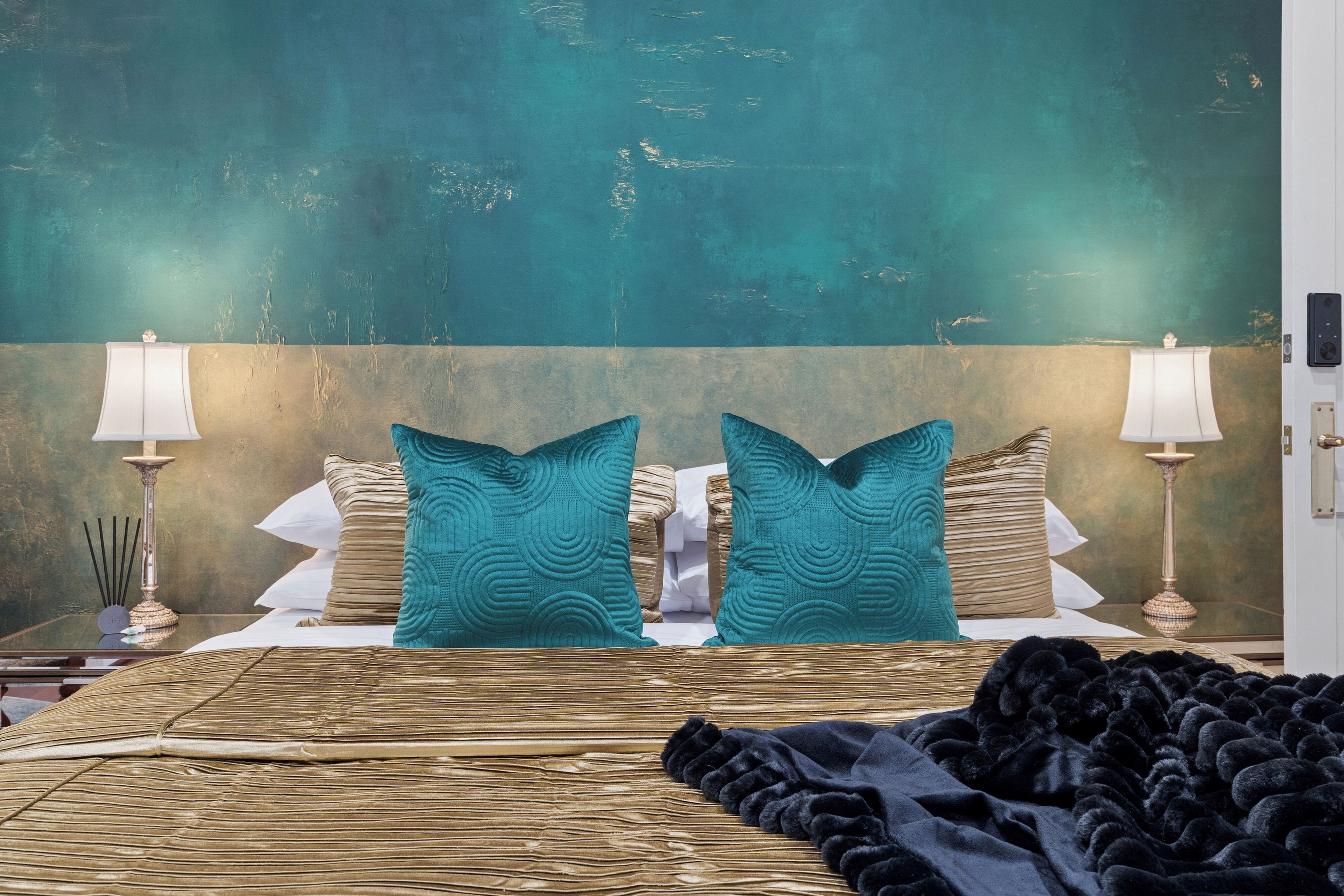 Una cama con cabecera, dos lámparas de noche y un almohadón grande color turquesa.