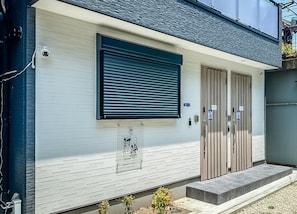 A building with a closed shutter, a security camera, and a sign indicating the building's name. - HANASTAY MUKAI MIGIWA & SUZU (Osaka)