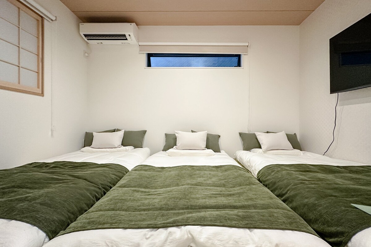 A room with three single beds, each with a green blanket and white pillows, a small window with a blue view, and an air conditioning unit on the wall.