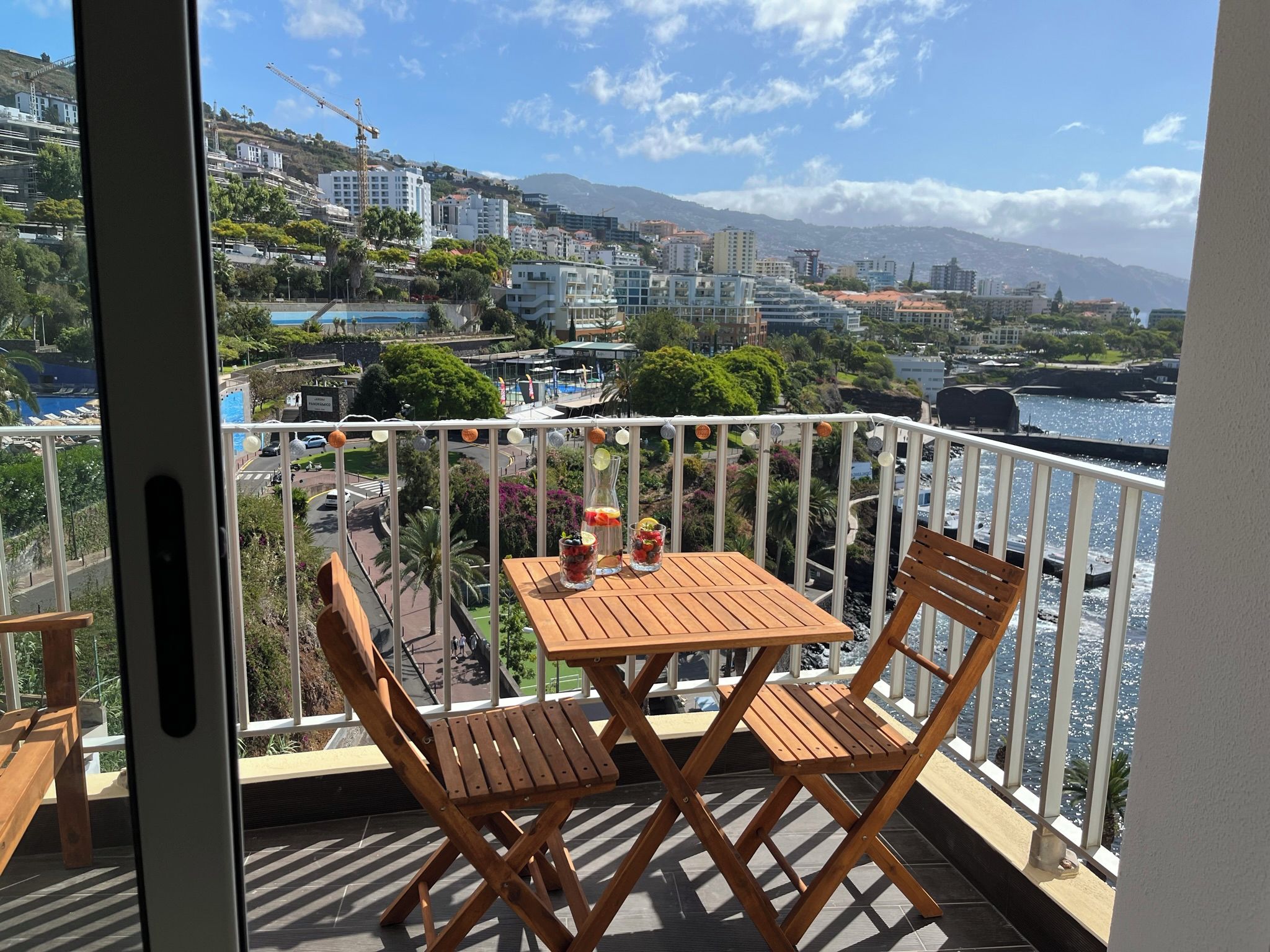 A balcony with a wooden table and chairs, overlooking a waterfront with buildings and a mountain in the background.