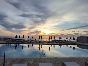 Une piscine bordée de chaises longues et d’ombrelles, où se reflète le coucher de soleil sur l’océan.