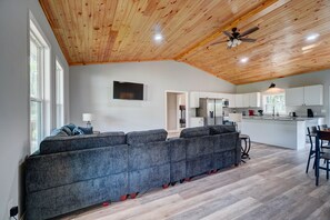 A modern living room with a large grey sectional sofa, a ceiling fan, and a kitchen area with white cabinets and a central island. - Peaceful New 3BR East Tennessee Retreat- Fire Pit - Deck- Garage (Sweetwater)