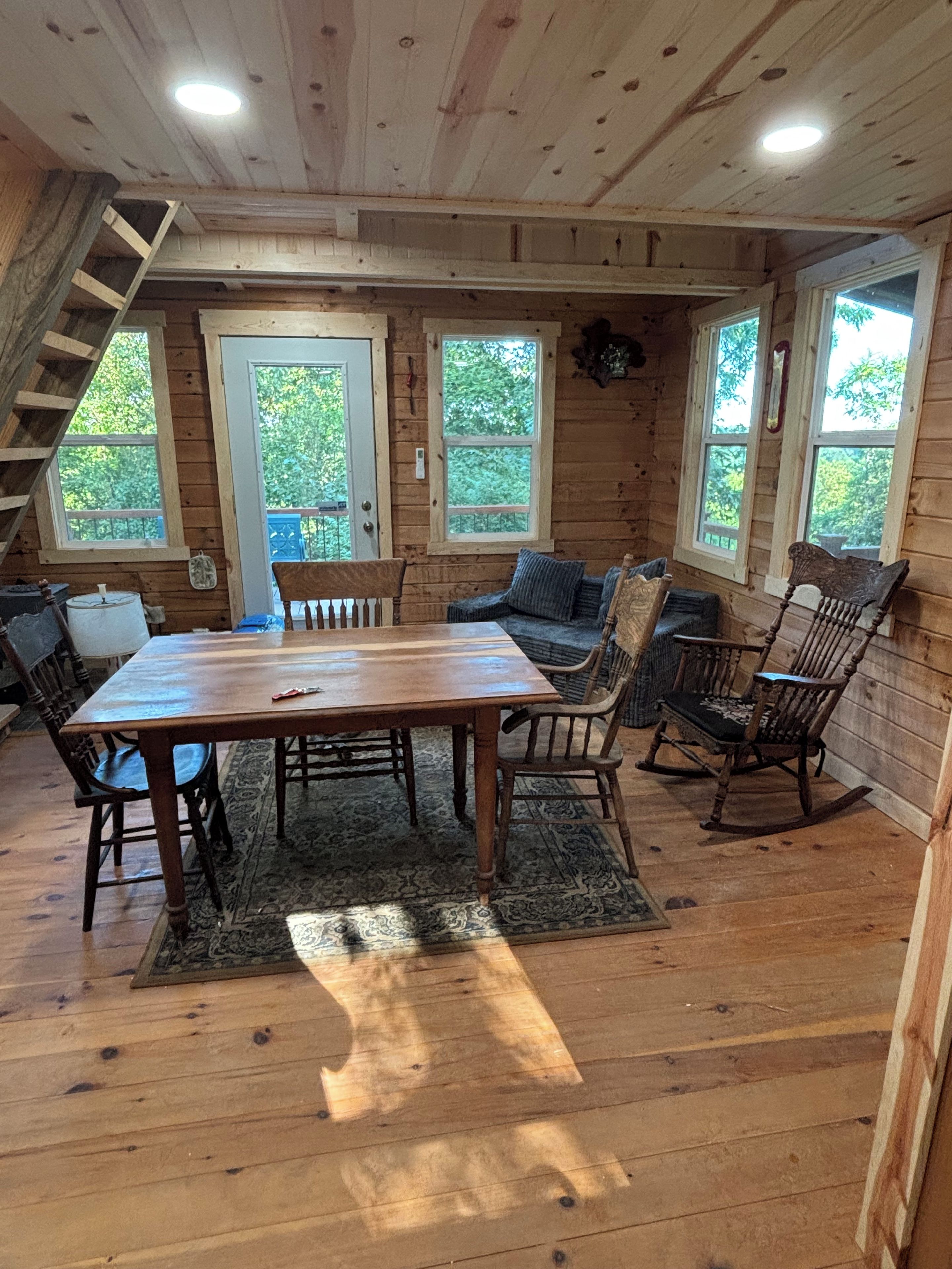 Een houten blokhut van binnen met een eettafel, stoelen, een bank, schommelstoelen, een trap en grote ramen.