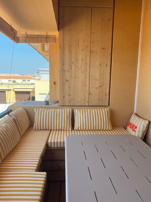 A sunlit balcony with a wooden door, a cushioned seating area with striped cushions, and a table with a white surface. - The prettiest studio apartment in Sète! Right on the beach, with views of the sea, the Pyrenees, and the sunset (Sète)