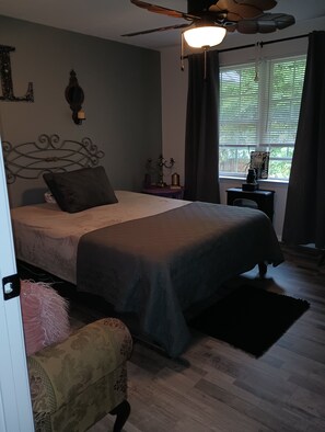 A bedroom with a bed, a ceiling fan, a bedside table with a lamp, a window with curtains, and a small table with decorative items. - 3-bedroom Home Away From Home.  More than a rental — it's an experience. (Annapolis)