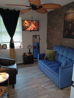A living room with a blue sofa, a ceiling fan, a television, and a wall with a poster. - 3-bedroom Home Away From Home.  More than a rental — it's an experience. (Annapolis)