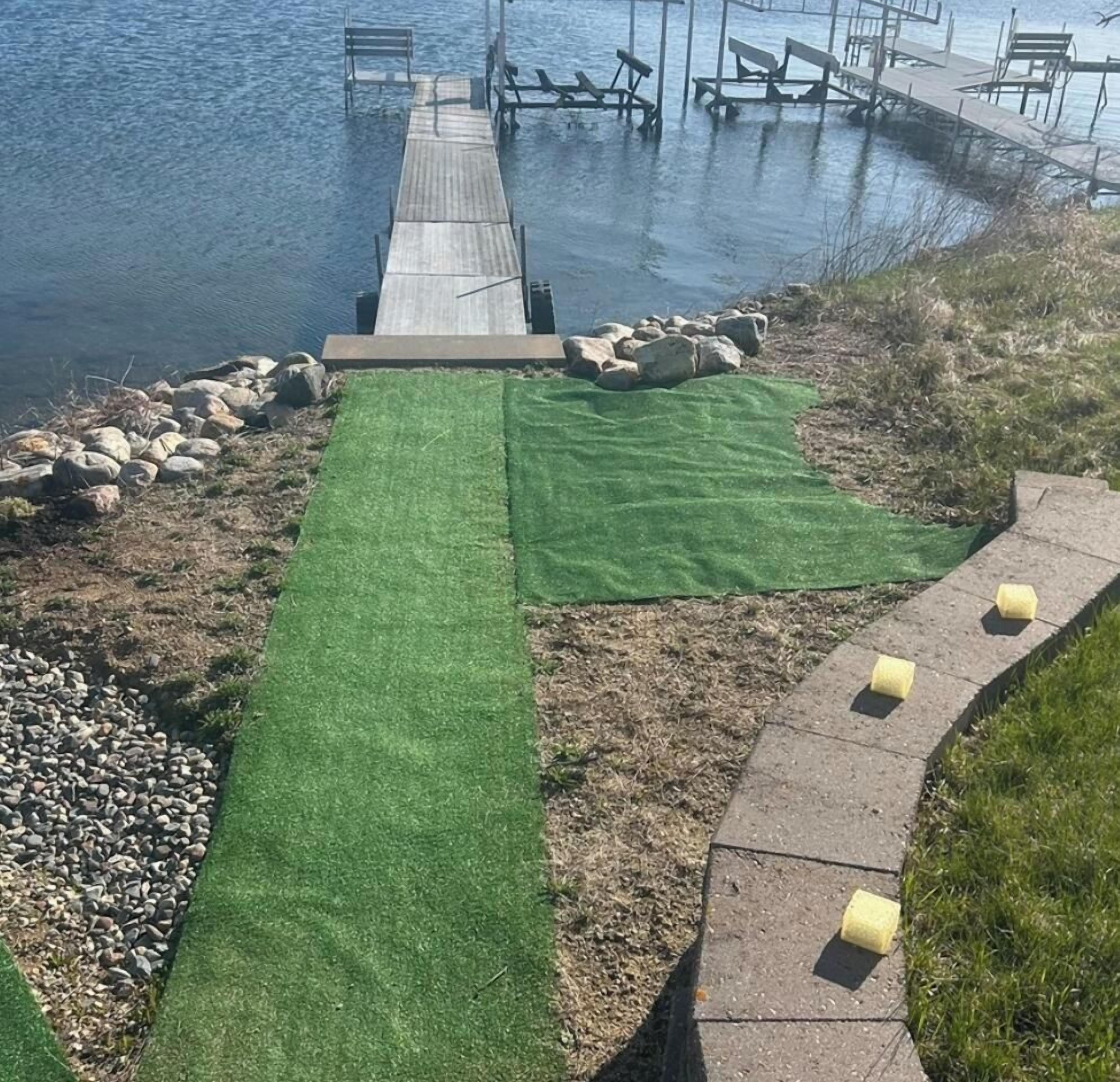 Una propiedad frente al agua con un muelle de madera que se adentra en el agua, un área verde con un camino y un borde de piedra.