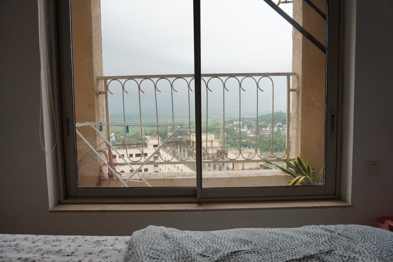 Ein Zimmer mit einem Fenster, das auf einen Balkon mit Metallgeländer und einen Blick auf Gebäude und Grünflächen blickt, ein Bett mit einem gemusterten Bettbezug.