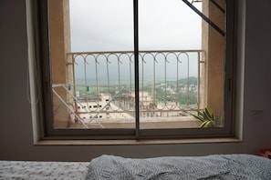 Ein Zimmer mit einem Fenster, das auf einen Balkon mit Metallgeländer und einen Blick auf Gebäude und Grünflächen blickt, ein Bett mit einem gemusterten Bettbezug.