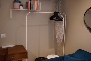 Ein Zimmer mit einem weißen Metallregal, auf dem Bücher und eine kleine Pflanze stehen, einem Nachttisch aus Holz mit einer Schublade, einem blauen Sofa und einem weißen Duschvorhang mit geometrischem Muster.