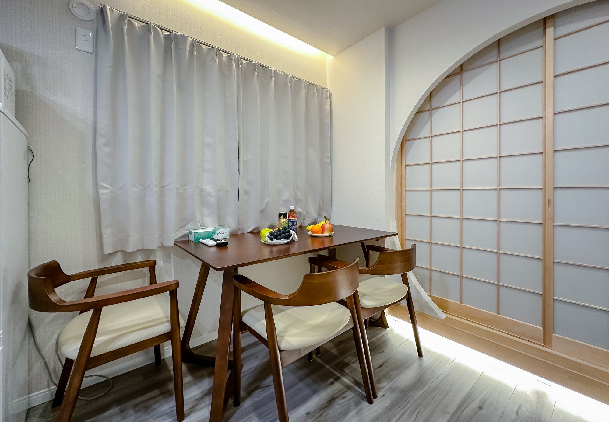 Una habitación con una mesa de madera, dos sillas, una mesita con frutas y una botella, una cortina y un panel shoji.