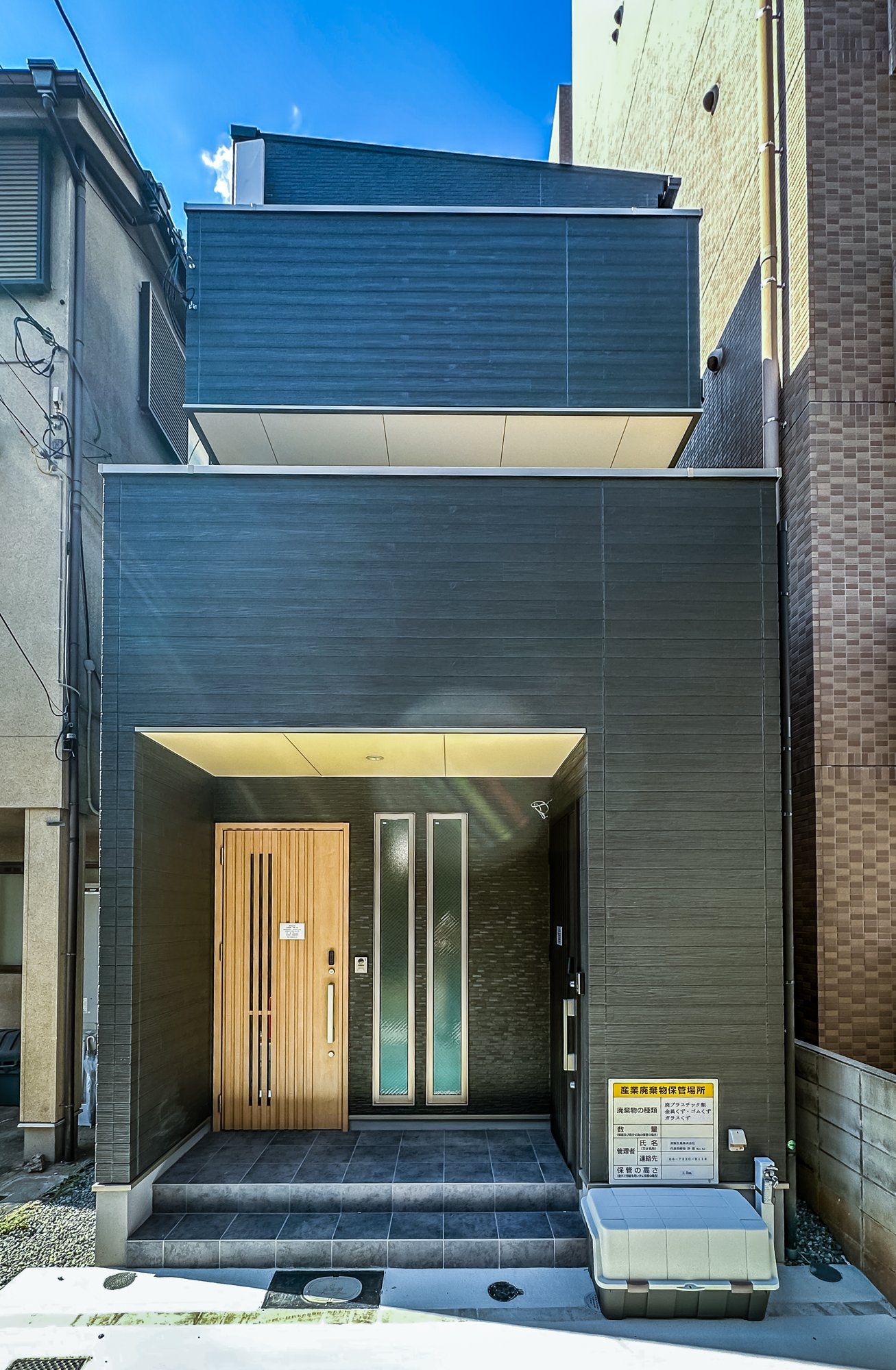 A modern building entrance with a dark facade, a wooden door, and a small step leading to the entrance.