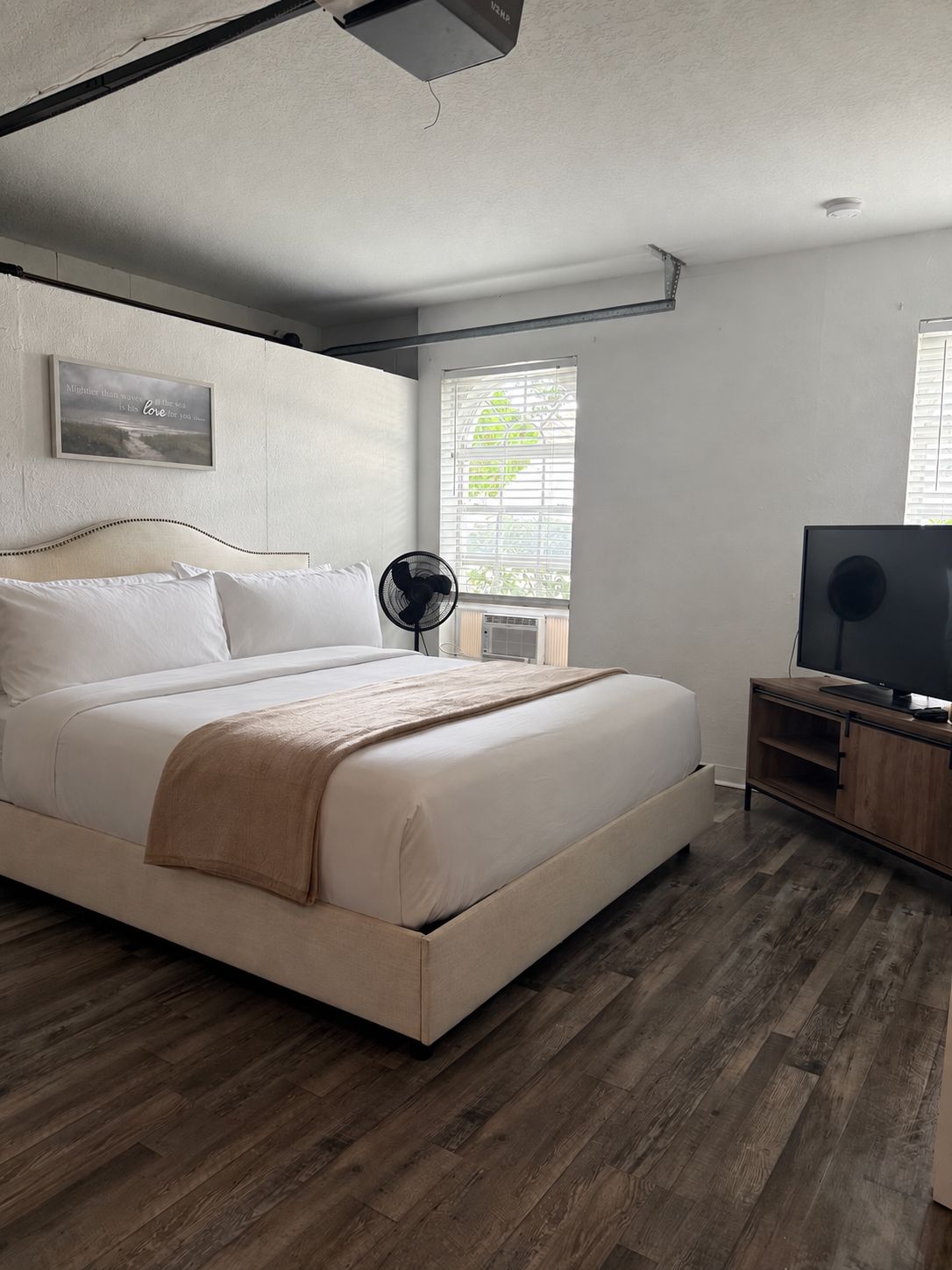 A bedroom with a bed, a TV, a fan, and a window with blinds.