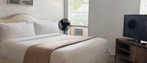 A bedroom with a bed, a TV, a fan, and a window with blinds.
