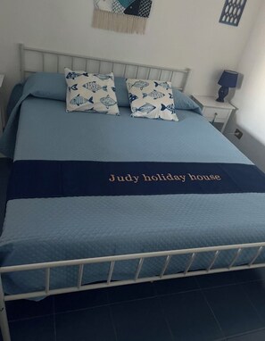 A bed with a quilted blue bedspread, two pillows with a fish pattern, and a dark blue bed runner with text. - Judy Holiday House (Sicilia)