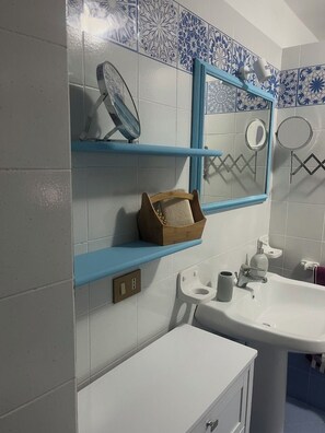 A bathroom with a white sink, a mirror, and a blue-framed shelf with a wooden basket. - Judy Holiday House (Sicilia)