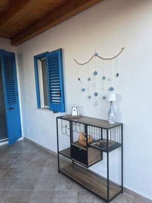 A room with a wooden ceiling, a window with blue shutters, a wall-mounted decoration with hanging items, a small table with a lamp, and a tiled floor. - Judy Holiday House (Sicilia)
