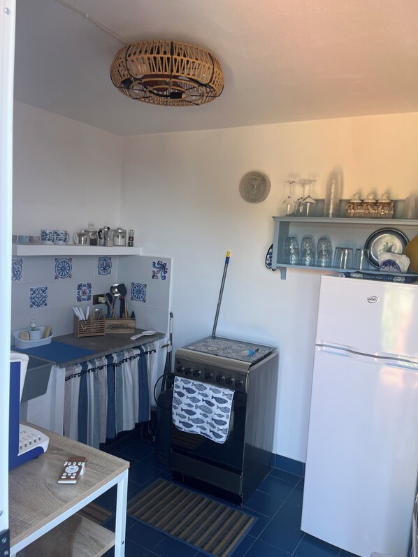A compact kitchen with a stove, oven, and refrigerator. There is a shelf with glassware and a hanging light fixture.