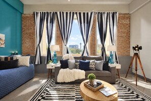 A modern living room with a blue couch, a grey sofa, and a wooden coffee table. There are two lamps on side tables, a tripod lamp, and a view of the cityscape through large windows with striped curtains. - The Bentley Collection - The Turquoise Loft (Dallas)