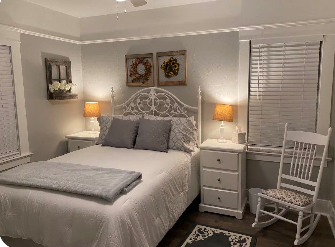 Quarto com cama branca, duas mesas de cabeceira com abajures, uma cadeira de balanço e enfeites nas paredes.