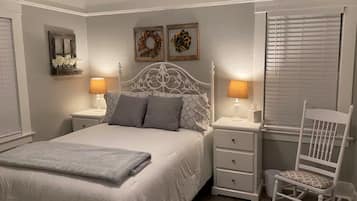 Quarto com cama branca, duas mesas de cabeceira com abajures, uma cadeira de balanço e enfeites nas paredes.